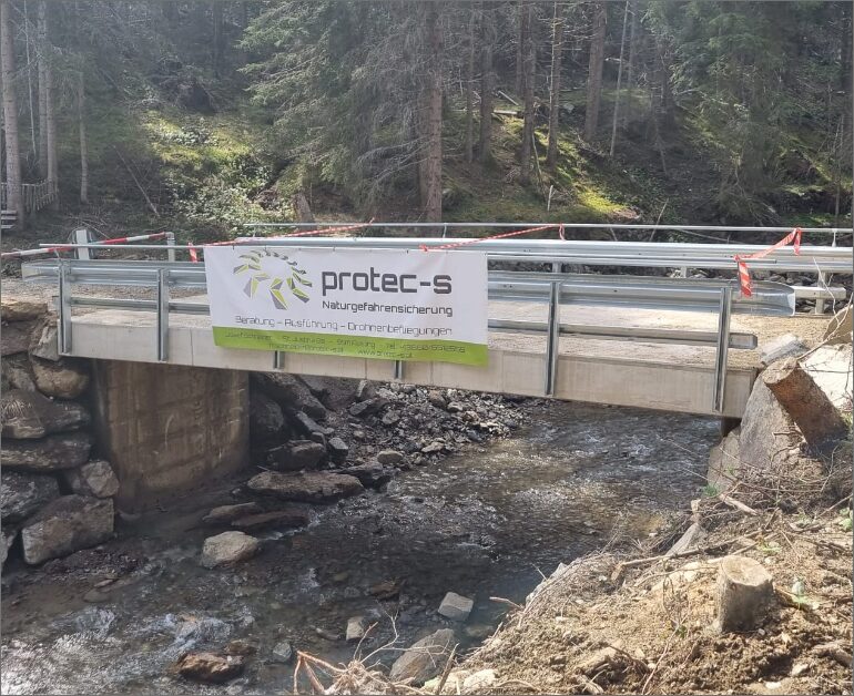 Eine im Bau befindliche Brücke über einen kleinen Bach, umgeben von einem Wald. Auf der Brücke ist ein Banner von „protec-s“ zu sehen.